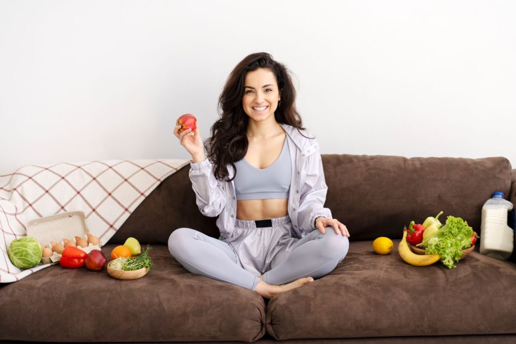 beautiful woman holding apple surrounded by healthy food on sofa